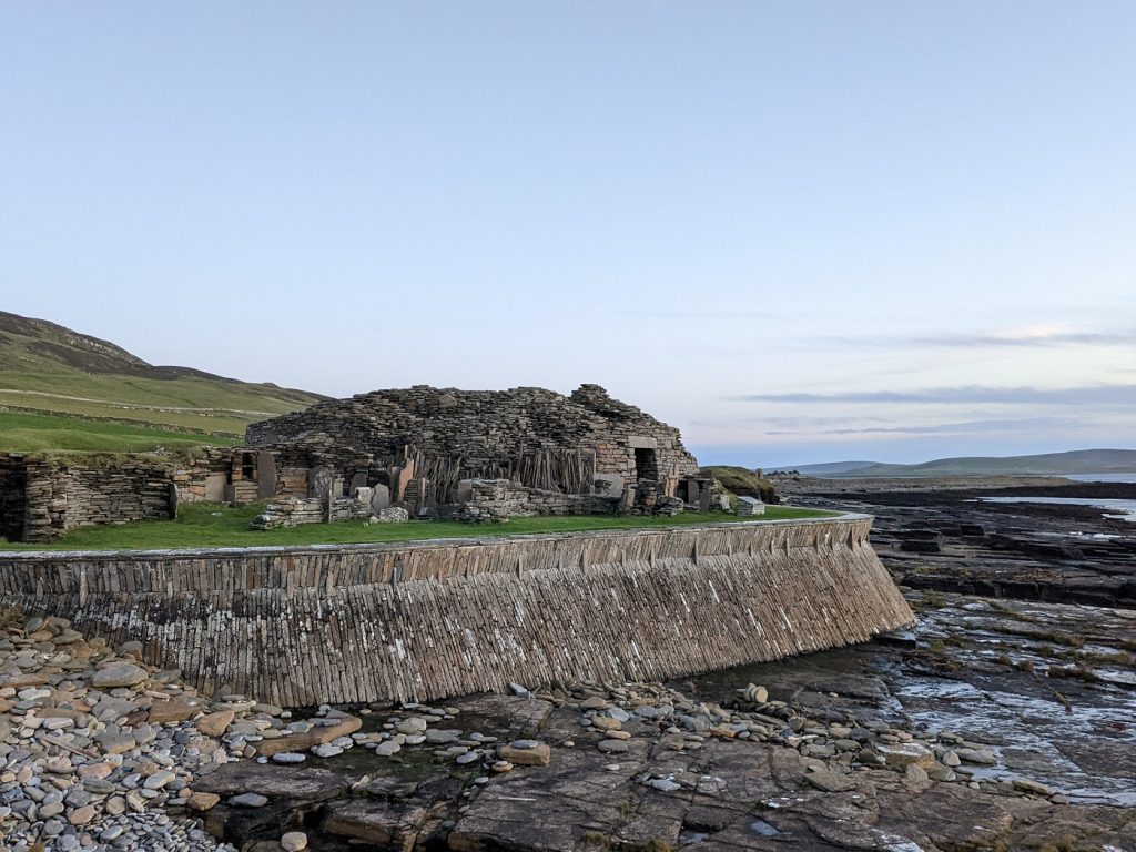 A ruinous round stone tower surrounded by a complex of smaller structures and stone partitions sits atop a wall-like stone barrier protecting it from the sea. A rocky shoreline is below the wall, and views extend across the water to neighbouring islands.
