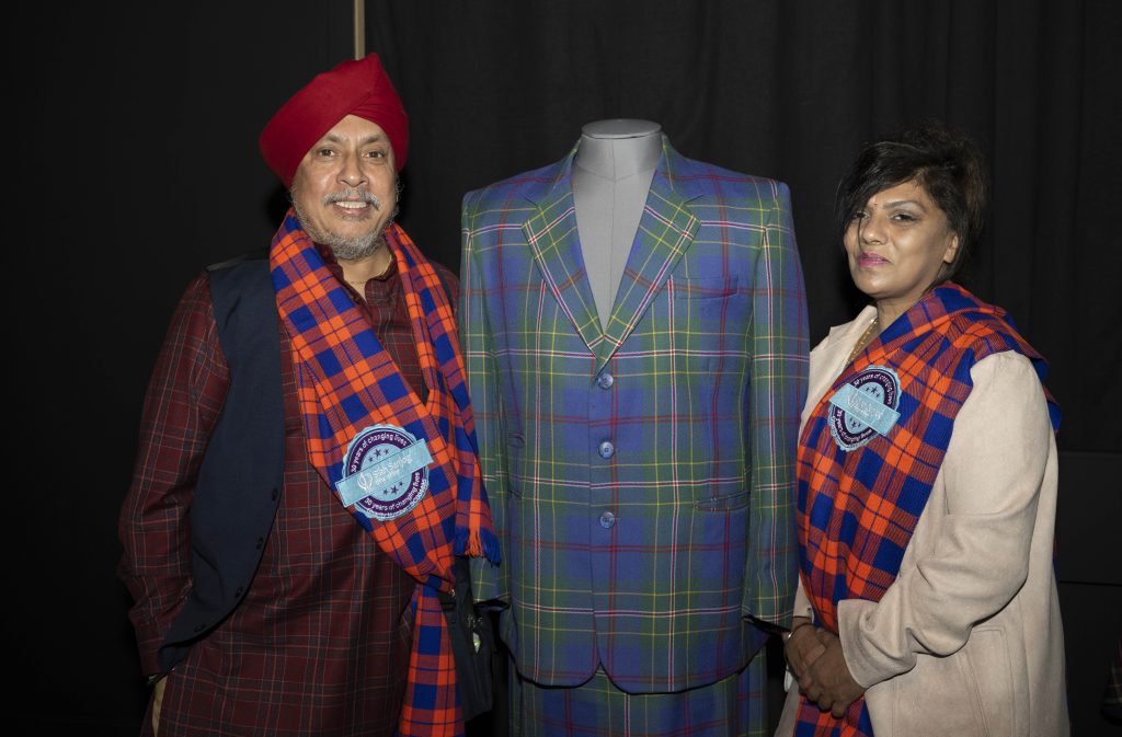 Two Members of Sikh Sanjog, one male and one female, wearing tartan sashes and stood with a tartan jacket in between them.