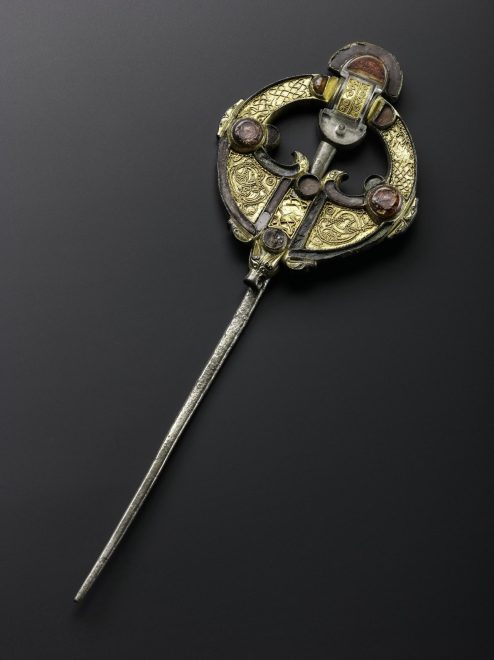 A shining brooch with a long silver pin and disc-shaped top against a dark grey background. The top is decorated with reddish gemstones, and its bronze surface is heavily decorated with swirling patterns.