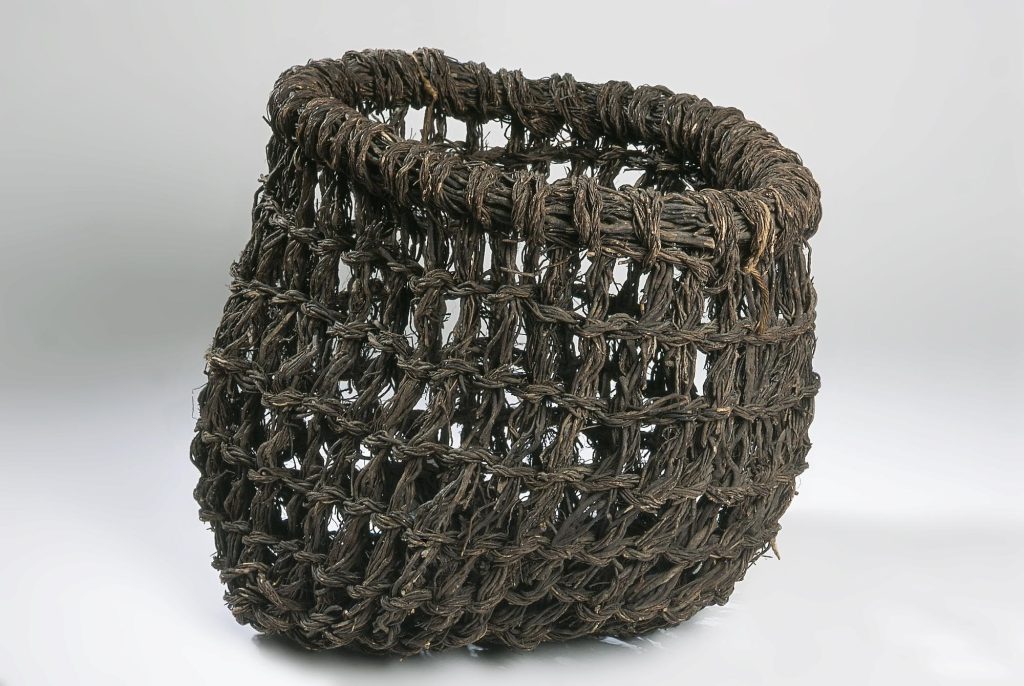 A basket of thick woven strands, turned greyish-brown with age and use, against a white background. Deeper than it is wide, the basket looks very sturdy.
