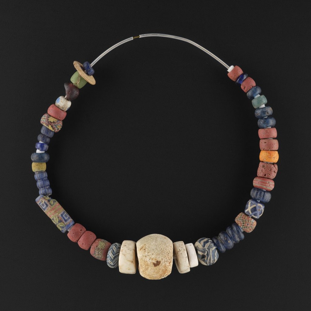 A very colourful and intricate string of beads, perhaps a necklace, of dazzling variety on a black background. The beads vary from small red, blue, and yellow discs to cylinders with multicoloured geometric patterns and a large central bead made of off-white bone.