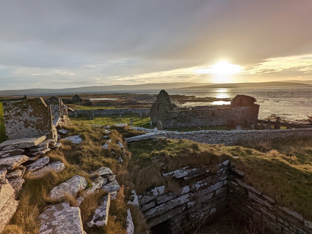 A radiant yellow sun sets across a channel of water behind low, distant hills. Perched atop a roofless square stone structure, the view extends over a ruinous church and a cluster of grey stone buildings along the shoreline.