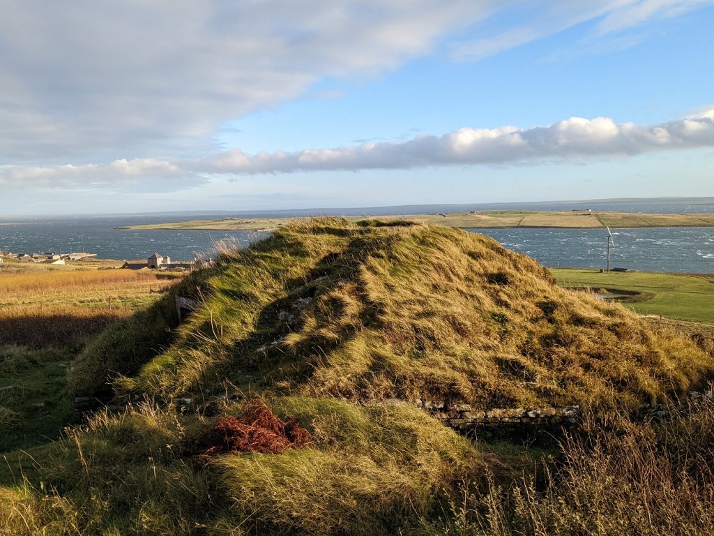 A one-storey mound covered in grass rises up from an area of long grass and heather. Behind it is an expansive sea-scape with several distant islands, and some farm buildings and wind turbines on the nearest shore.