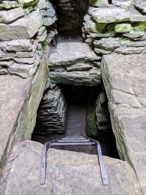 The upper level of an ancient stone chamber. The camera looks down a square opening with an iron ladder leading down to the lower level. Piles of grey worked stone flank the opening, which leads into darkness.