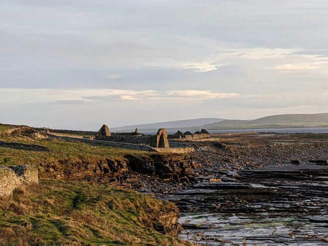 Several ruinous stone buildings, one after the other, look out over a raised shoreline strewn with seaweed. The ruins of a medieval church stand within a low stone wall. Low, rolling hills are visible across a channel of water.