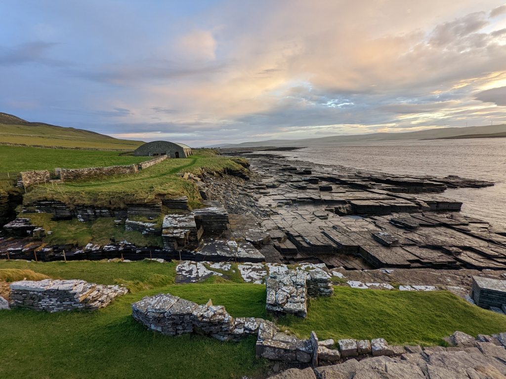 Under broad, cloudy skies turning colourful at sunset is a stony shoreline with several stone structures built near the edge. Rectangular flagstones slope down into the waters, and a rock trench cuts of a promontory in the foreground. A faint path leads to a hangar-like structure in the middle distance. 