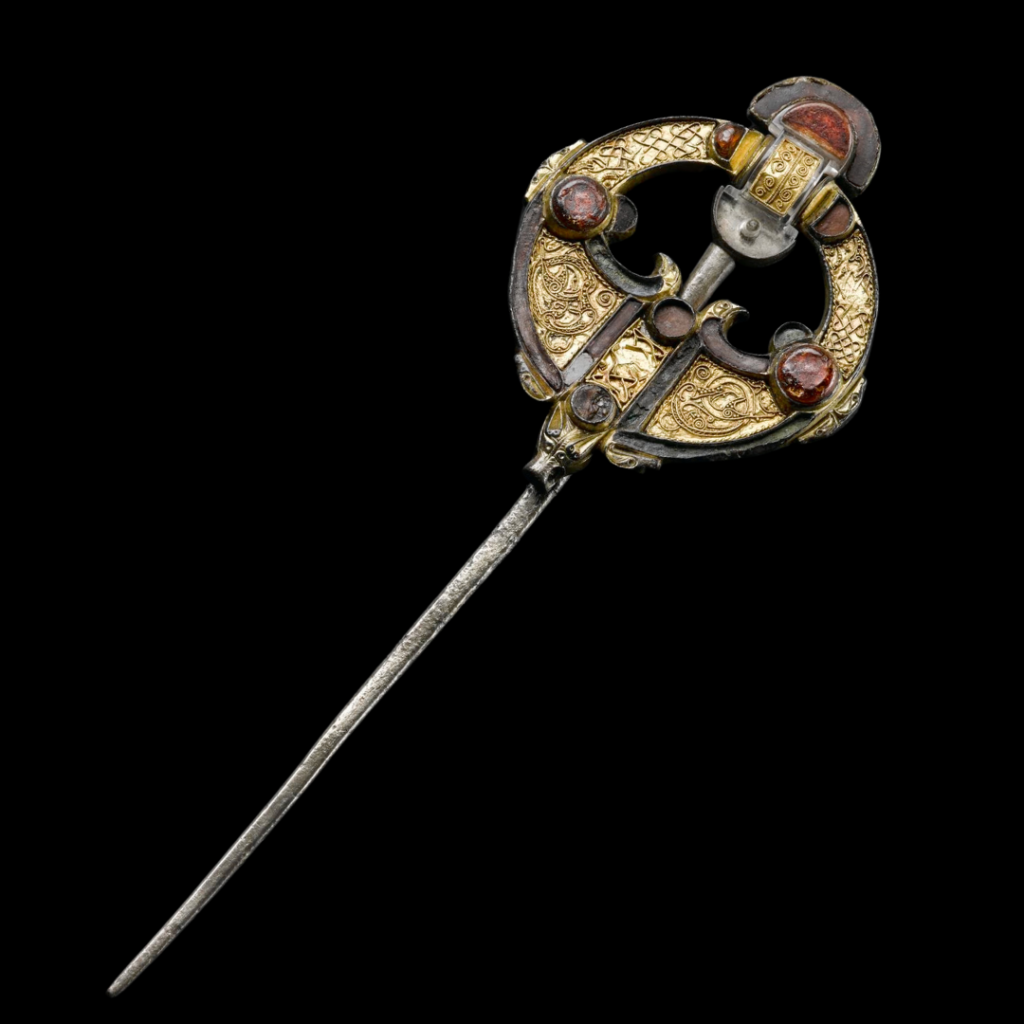 A shining brooch with a long silver pin and disc-shaped top against a dark grey background. The top is decorated with reddish gemstones, and its bronze surface is heavily decorated with swirling patterns.