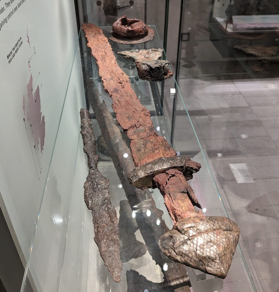 A glass museum case displays several Viking-era weapons, all badly rusted with rough edges. A long sword with a decorated pommel takes up the most space, alongside an axehead, spearhead, and a shield boss in the shape of a dome.