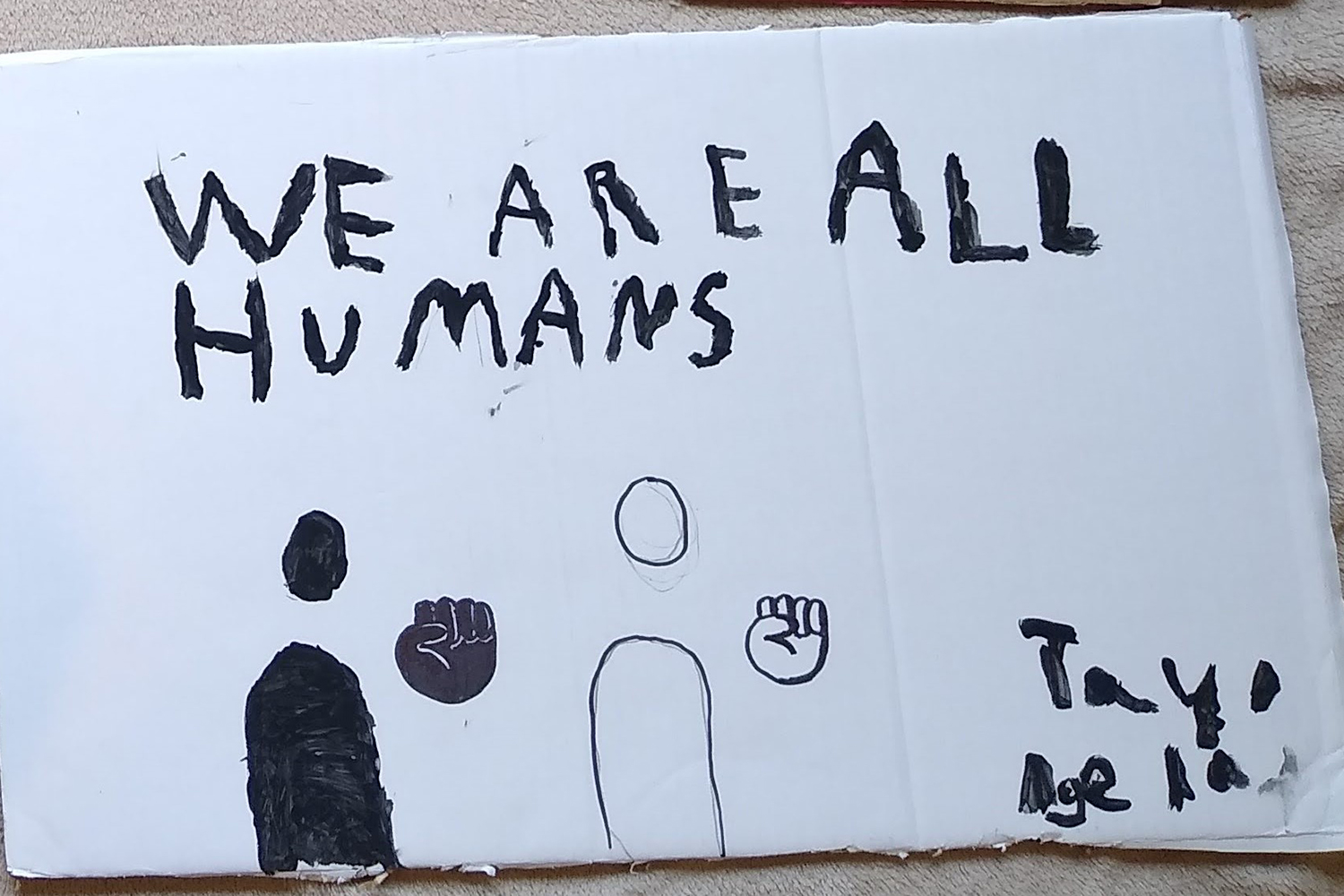 White cardboard placard with images and text drawn in black ink. It reads We are all humans, Tayo Age 10.