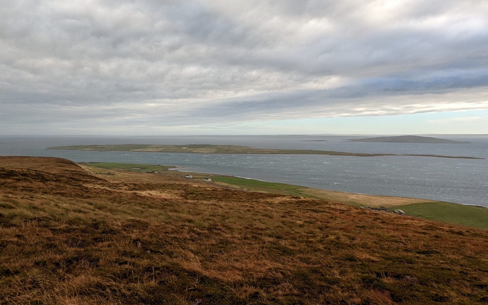 Standing in yellow and brown moor grass on a hillside, a very thin and low-lying island is in the middle of an expansive body of water in the distance. Several other low islands rise up beyond it, with grey cloud cover above.