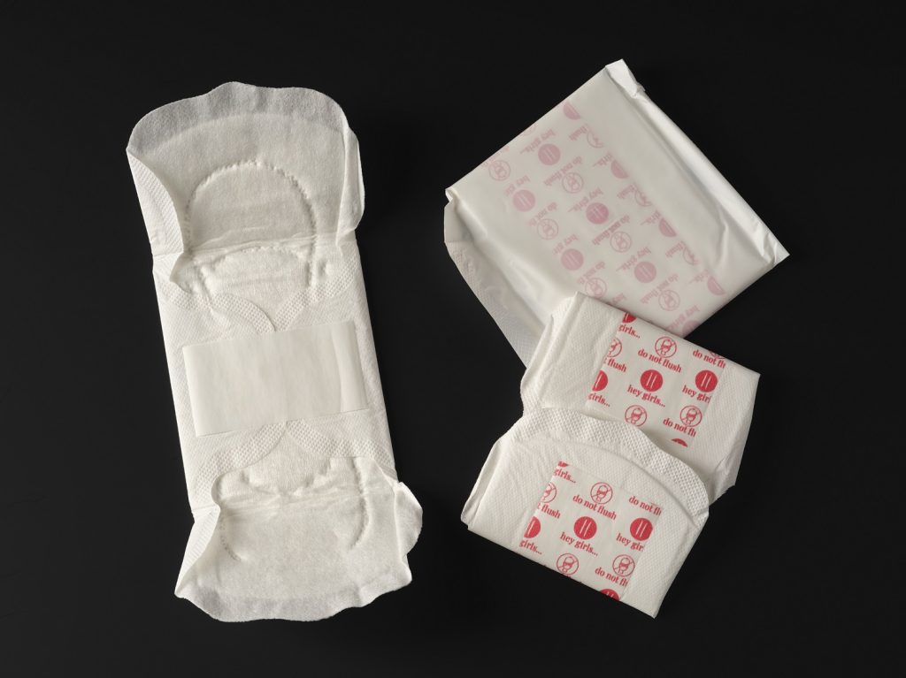 A white, unfolded period pad positioned vertically against a black background. To its right is a folded pad decorated with red instructions, and a sealed pad pack.