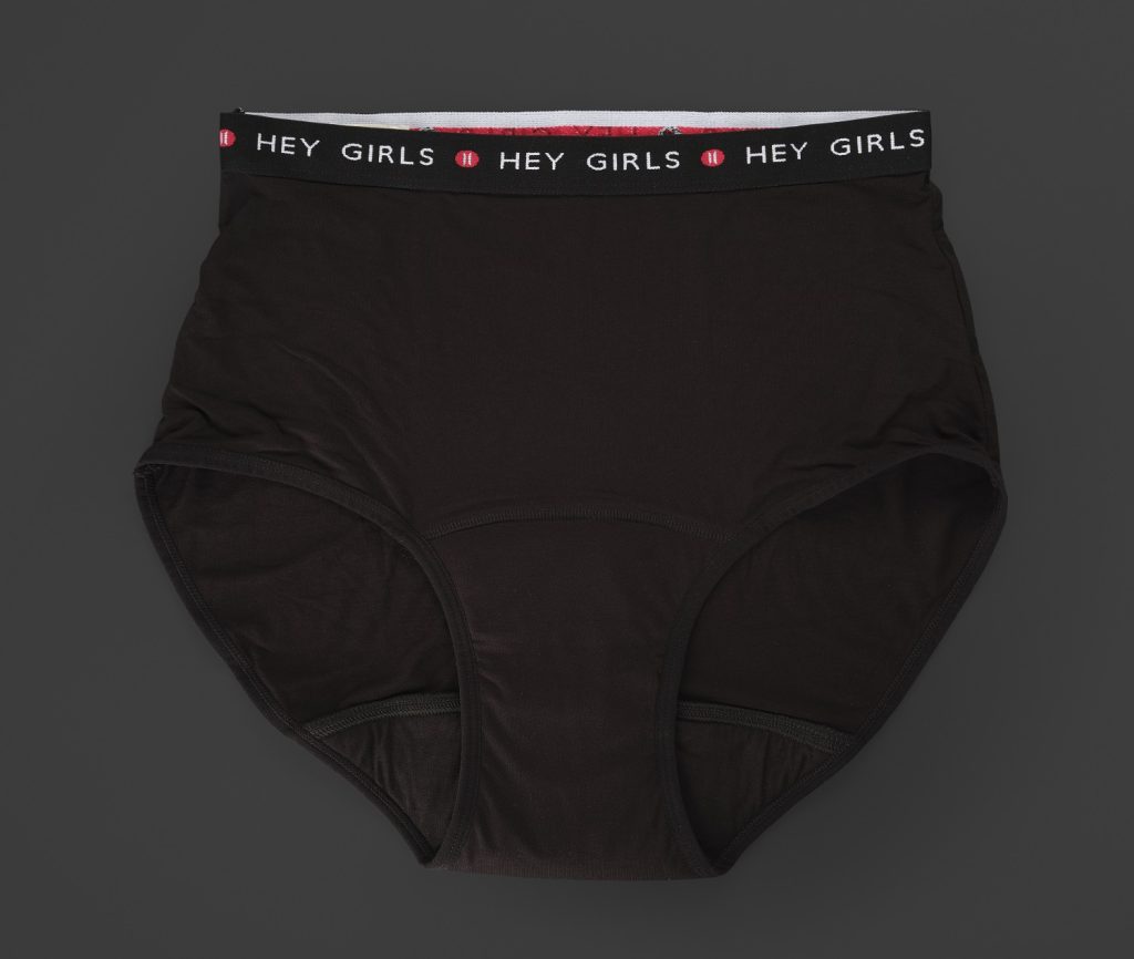 A pair of black briefs laid out flat on a dark grey background, with the words 'Hey Girls' repeated along the top band.