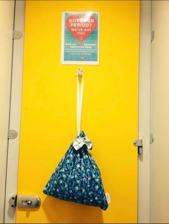 A blue canvas bag with a star pattern hangs from a metal hook on a tall, bright yellow toiler stall door.