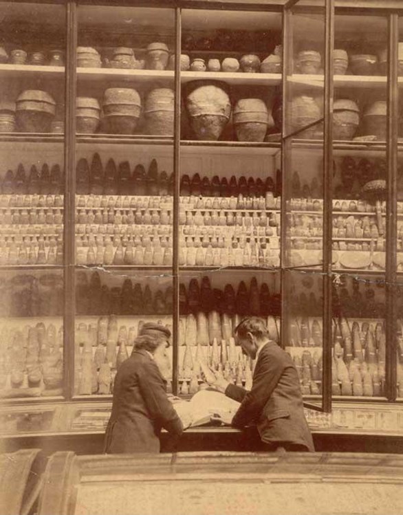 Sepia image of two people in suits examining a large open book on a desktop. Above them is a huge display behind glass panels of hundreds of ancient artefacts, including pottery and stone tools.