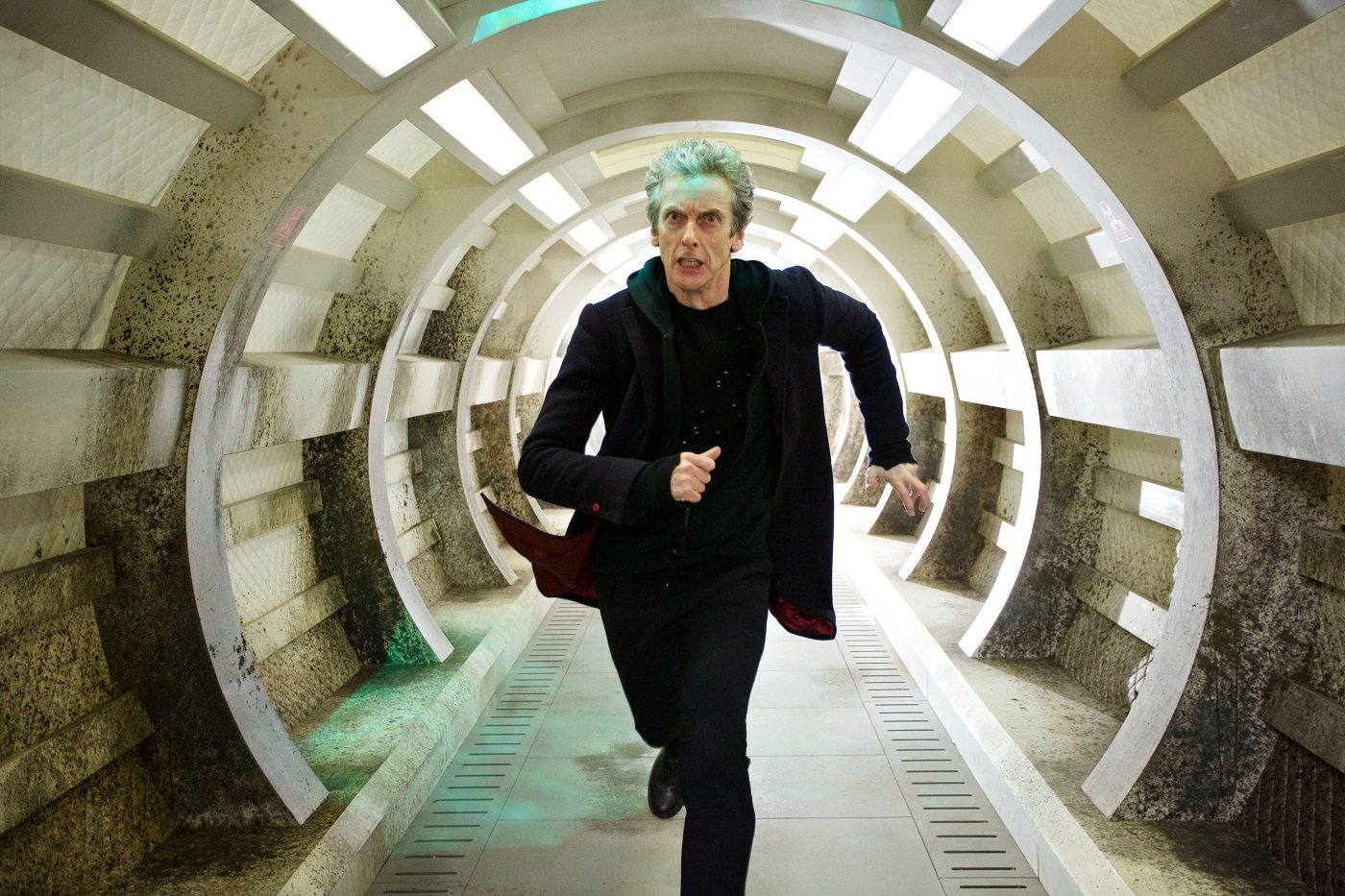 Peter Capaldi as Doctor Who running down a futuristic, circular corridor straight towards the camera. He's wearing an all-black outfit and looks worried.