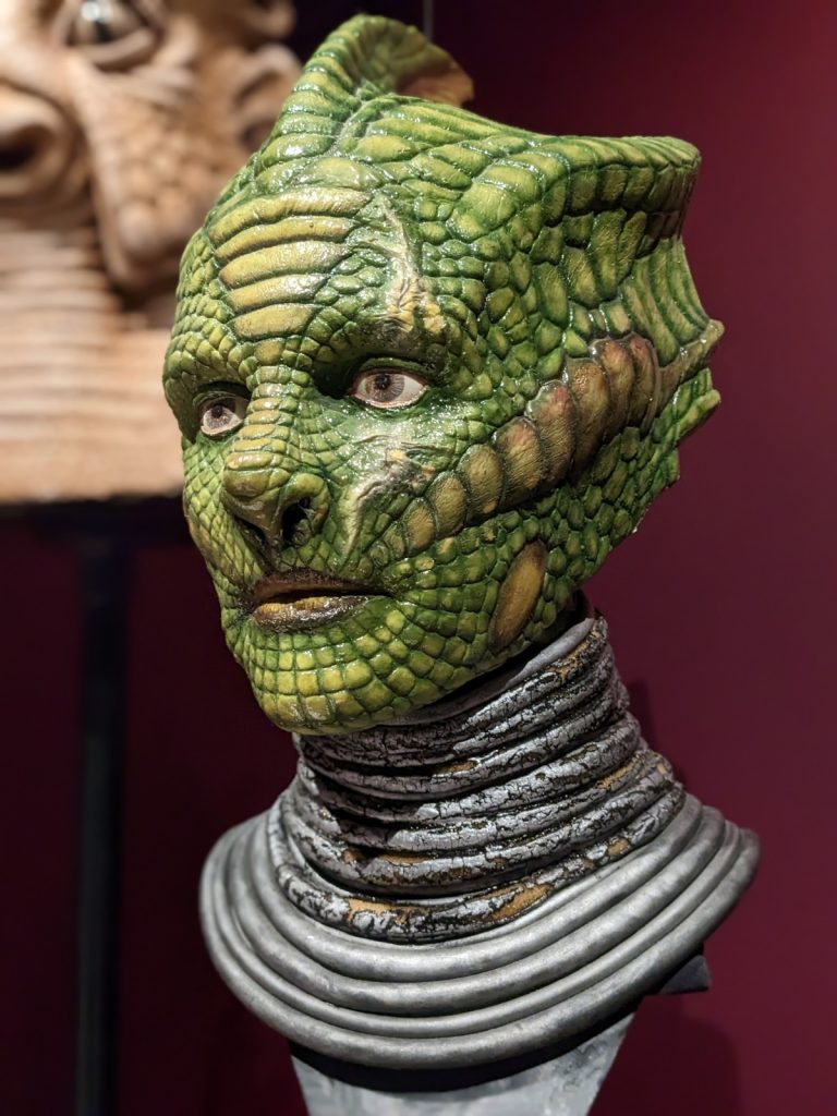 Closeup of a prop character's head mounted within a museum exhibition with dark red walls. The head is humanoid but green with scales like a lizard and several ridges creating an elongated skull. Though alien, the character looks intelligent and compassionate.