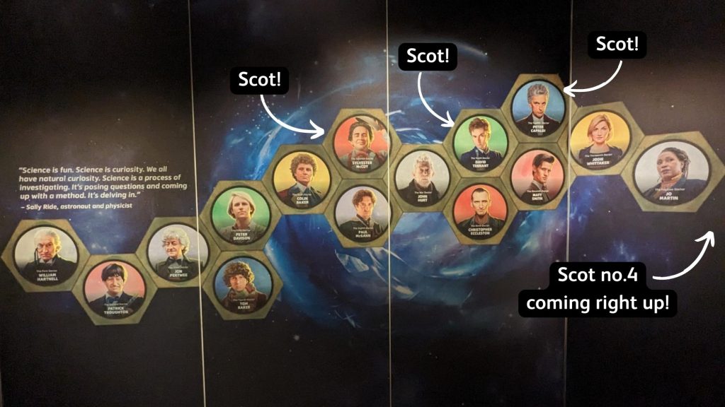 A graphic on the wall of a museum exhibition showing each of the actors who played Doctor Who. Each actor is shown from the chest up within a hexagon, with the hexagons forming a chain. A galaxy swirls on the black surface behind them.