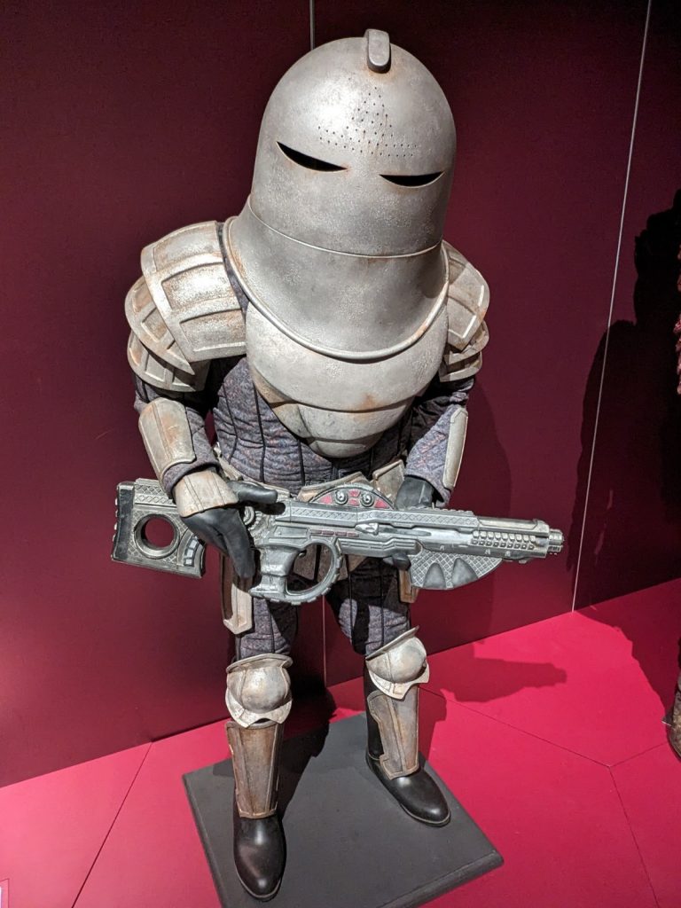 A suit of Sontaran armour within a museum exhibition with red walls. The armour is very stout and bulky, with a massive domed helmet resembling a 19th century diver's gear, thick steel shoulder pads, shin and wrist guards, and a futuristic grey metal gun.
