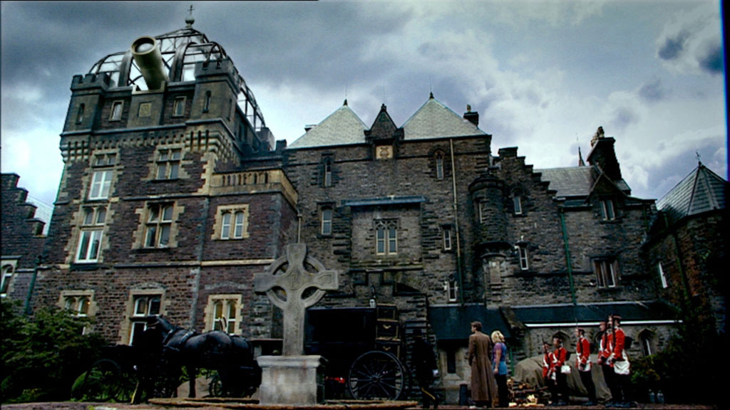 The Doctor and Rose approach a black horse-drawn carriage flanked by guards in red coats, all in front of a turreted mansion house with a stone Celtic cross in the foreground. A massive telescope lens resembling a cannon projects out from a steampunk-style addition on top of the mansion.