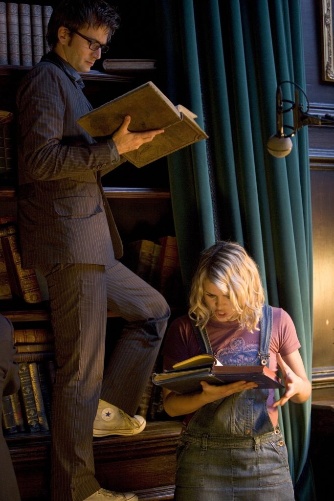 David Tennant and Billie Piper study books within a fancy library lined with navy blue curtains. Tennant looks neutral and is standing on part of a bookcase elevating him well above Piper, while Piper is reacting with excitement to something in the book she has.