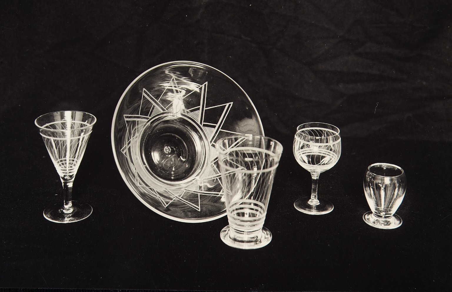 Five glass objects lined up against a plain black background. From left to right are a tumbler glass with geometric patterns, a large bowl with triangular decorations laid upright on its side, a cone-shaped glass with wavy lines, a goblet with wavy lines, and a small pear-shaped vessel that almost looks metallic.