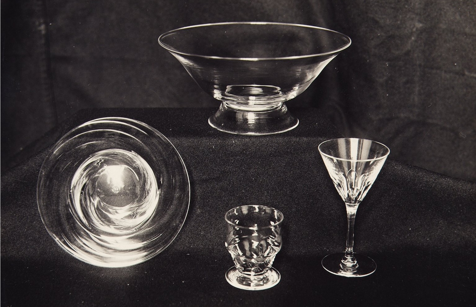 Black and white photo of a display case lined with dark cloth. Inside are four clear glass objects: form left to right, a wide bowl with swirling pattern, a clear wide bowl, a short and stout cup with textured ridges, and a wine glass.
