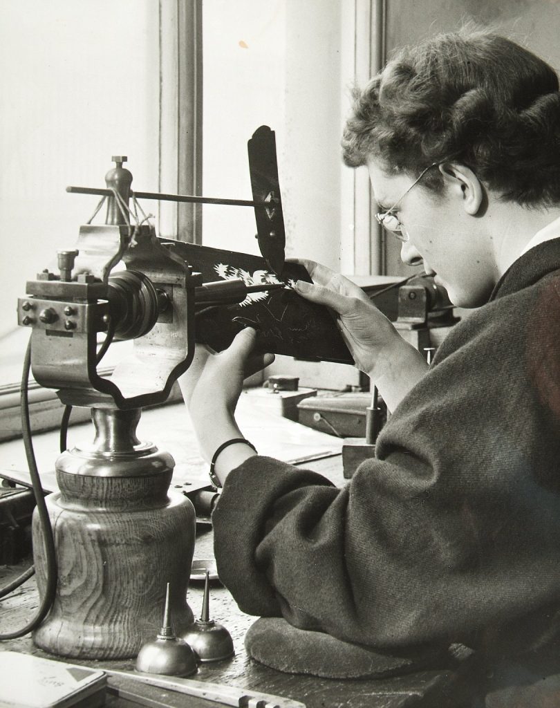 Black and white photo of a woman with short, wavy hair, glasses and a thick billowy blouse intently working on cutting out a pattern from a panel. She is using a large tool with wooden base and large metal clamp to hold the panel in place. She looks focused and proud.