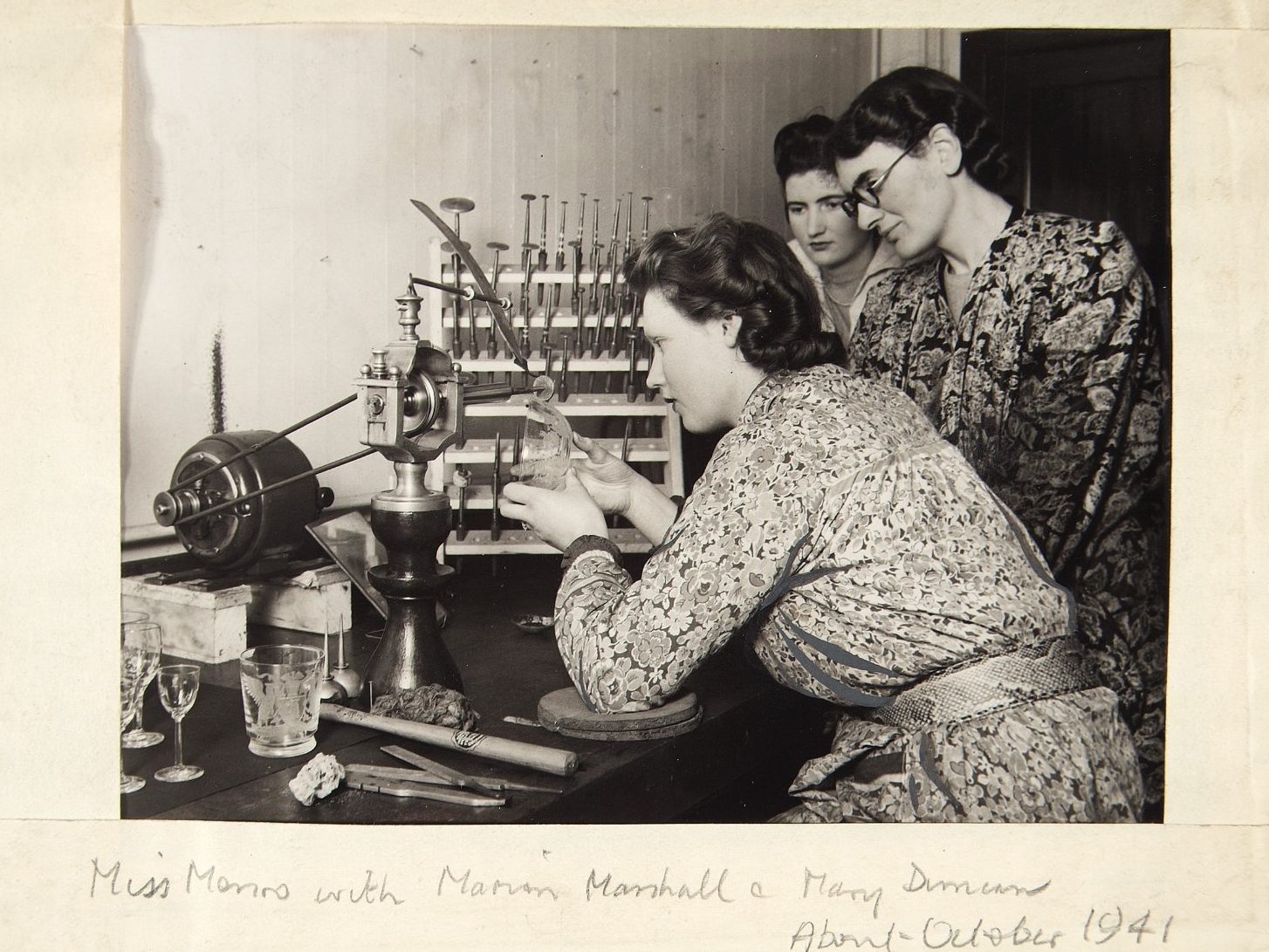 Black and white photo framed by off-white paper bearing pencil script at the bottom dating it to 1941. In the image, three women in floral dresses examine a glass object at a wooden table filled with various tools and glass objects.