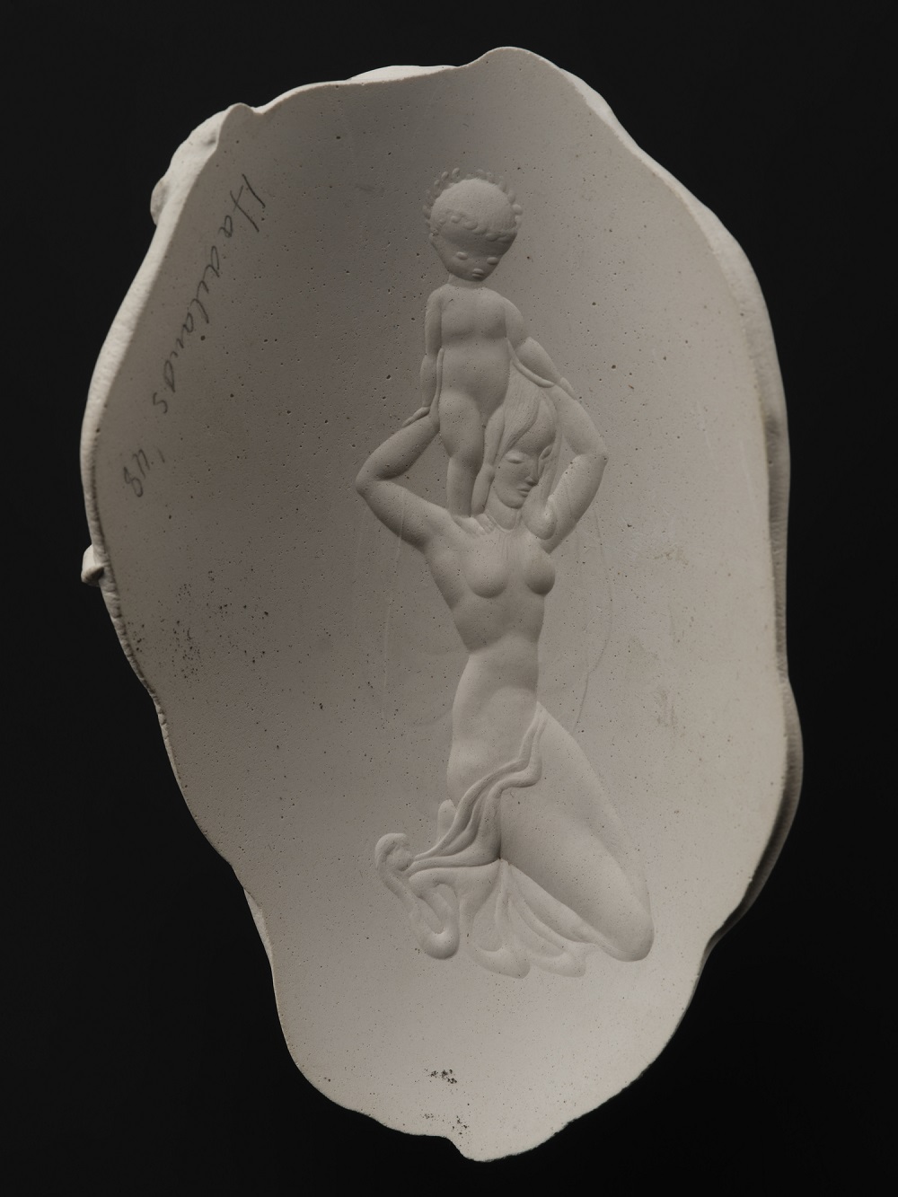 A white ceramic fragment shaped like an egg with wavy edges against a black background. On the white surface is a raised carving of a nude woman kneeling and holding a nude child on her shoulders. The child is wearing a laurel crown, and the woman has a cloth draped across her waist.