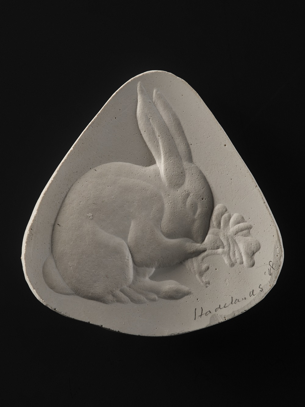 A white ceramic piece, triangle-shaped like a guitar pick, against a black background. A raised carving of a simplified rabbit nibbling on a leaf takes up most of the curved inner surface.