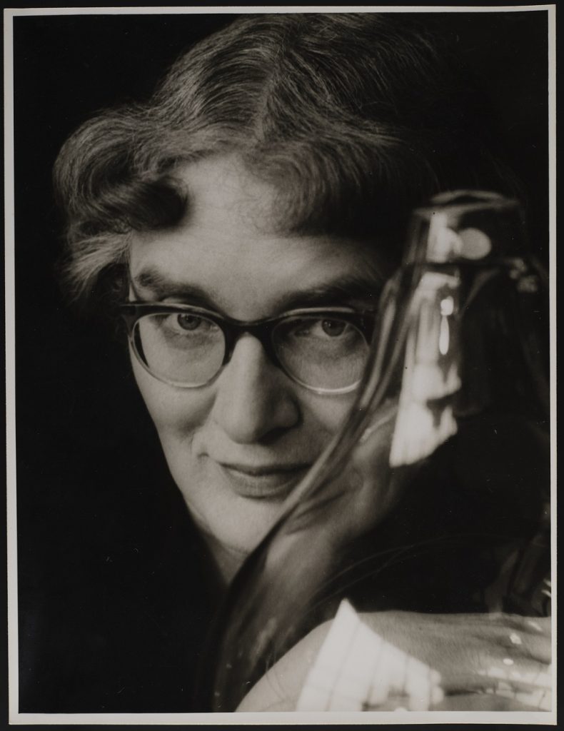 Closeup portrait of a woman with short, curly hair and thick glasses smiling beguilingly behind a bulb-shaped glass vessel. The portrait feels intimate and personal.