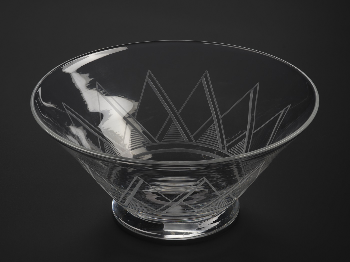 A clear glass bowl, much wider at the top than the bottom, decorated with a pattern of repeated triangles radiating up from the basin. On the left, part of the bowl catches a glare.