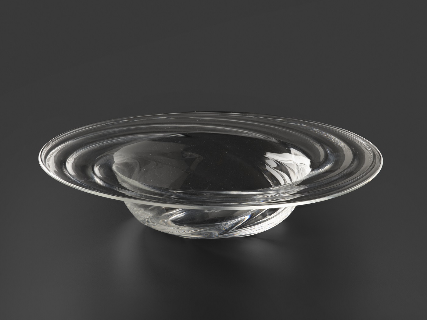 A translucent glass vessel shaped like an upside-down, broad-rimmed hat against a dark grey background. Lines on the glass rim resemble water spiraling in a sink.