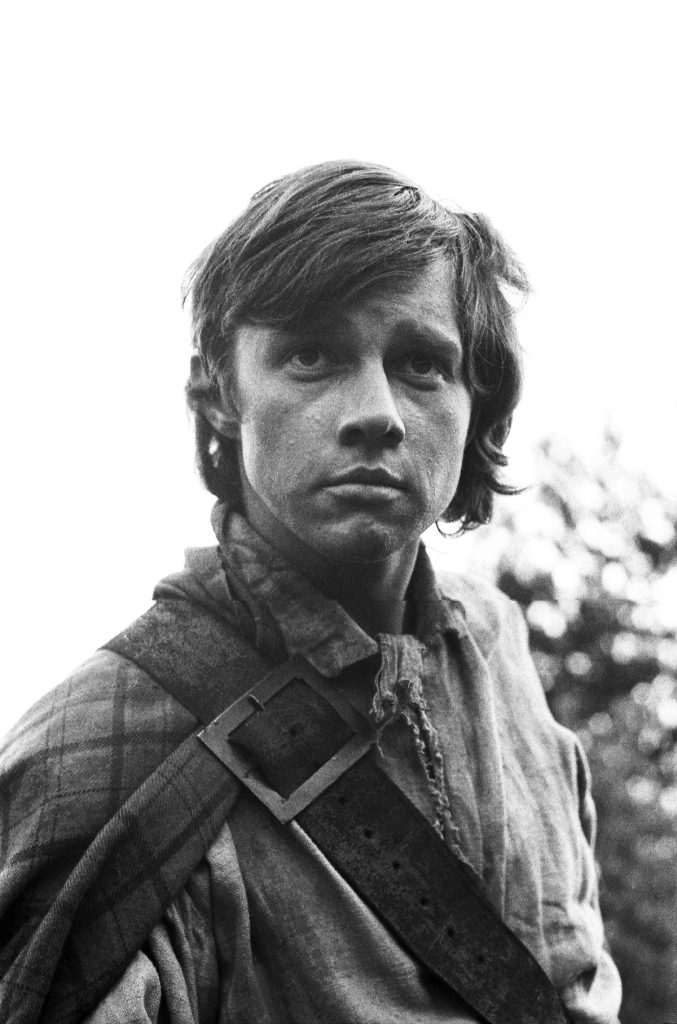Black and white image of a handsome young man vaguely resembling Luke Skywalker seen from the chest up, looking over the camera. He is dressed like a 18th century Highlander in a tartan cloak, over the shoulder strap, and discoloured white shirt. 