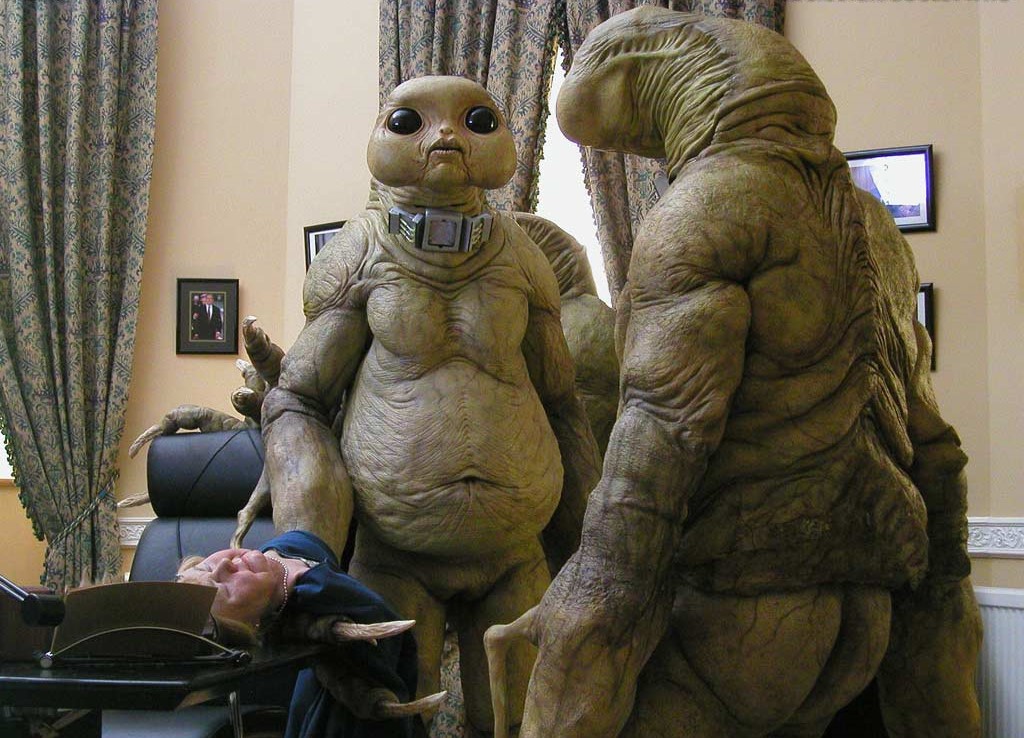 Scene from Doctor Who in which a trio of aliens, humanoid in form and light green-coloured with beady black eyes and loose, wrinkled skin, stand over an unconscious woman in a doctor or dentist's office.