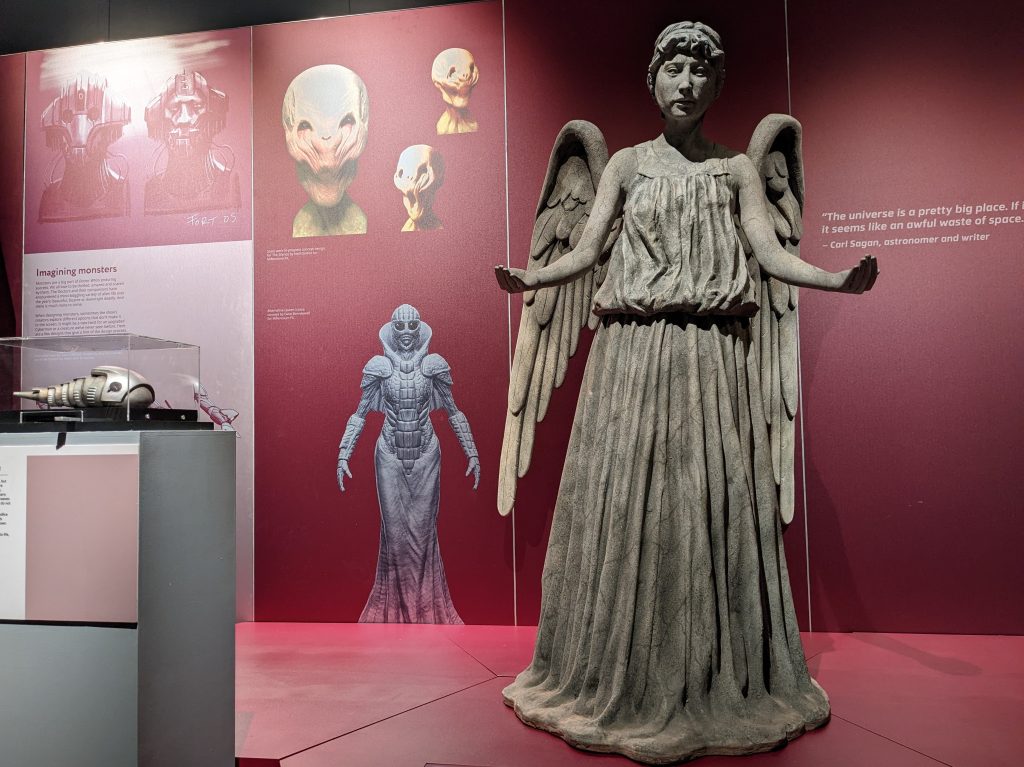 A museum display of red panels showing various scary aliens fronted by a statue of a Weeping Angel, a Doctor Who monster which takes the form of a stone angel like those found in cemeteries or cathedrals.