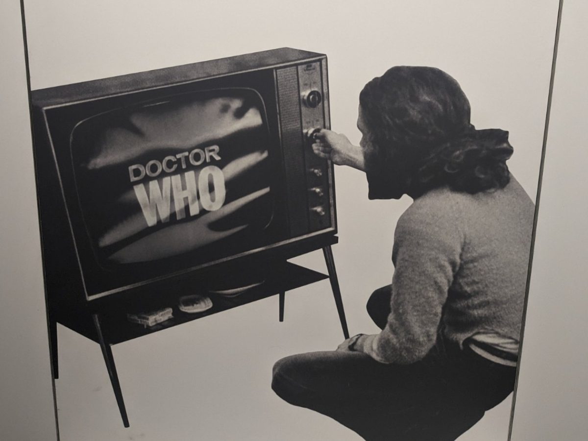 Panel in a museum exhibition showing a black and white image from the late 1960s or early 1970s. A man is seated cross-legged in front of a freestanding TV.