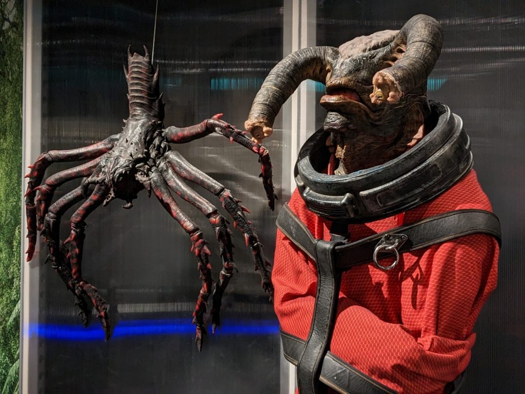 Two Doctor Who aliens side-by-side against a reflective grey background. On the left is a Dalek out of its shell, resembling a cross between a spider and a lobster. On the right is a Teller, a humanoid alien with two appendages on its head resembling an elephant's trunk. The Teller is constrained by an orange straight-jacket and several thick leather straps.