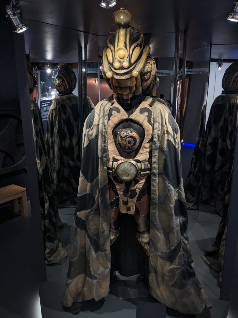 Costume for a Time Lord from Doctor Who, dimly lit and flanked by mirrors which show off each side of the costume. The costume looks like a cross between an astronaut and an insect, with bulbous helmet, camouflage-coloured cloak, and a utility belt.