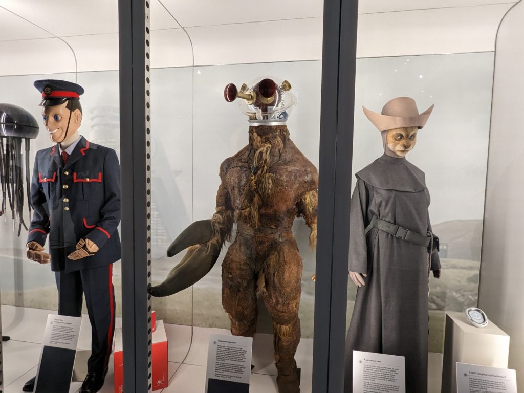 Three full-size Doctor Who costumes inside a brightly lit display case that resembles the inside of a sleek, white spaceship. From left to right are a humanoid robot with uncanny wide eyes, a creature that looks like a cross between a Yeti and a lobster, and a humanoid cat dressed in plain grey robes.