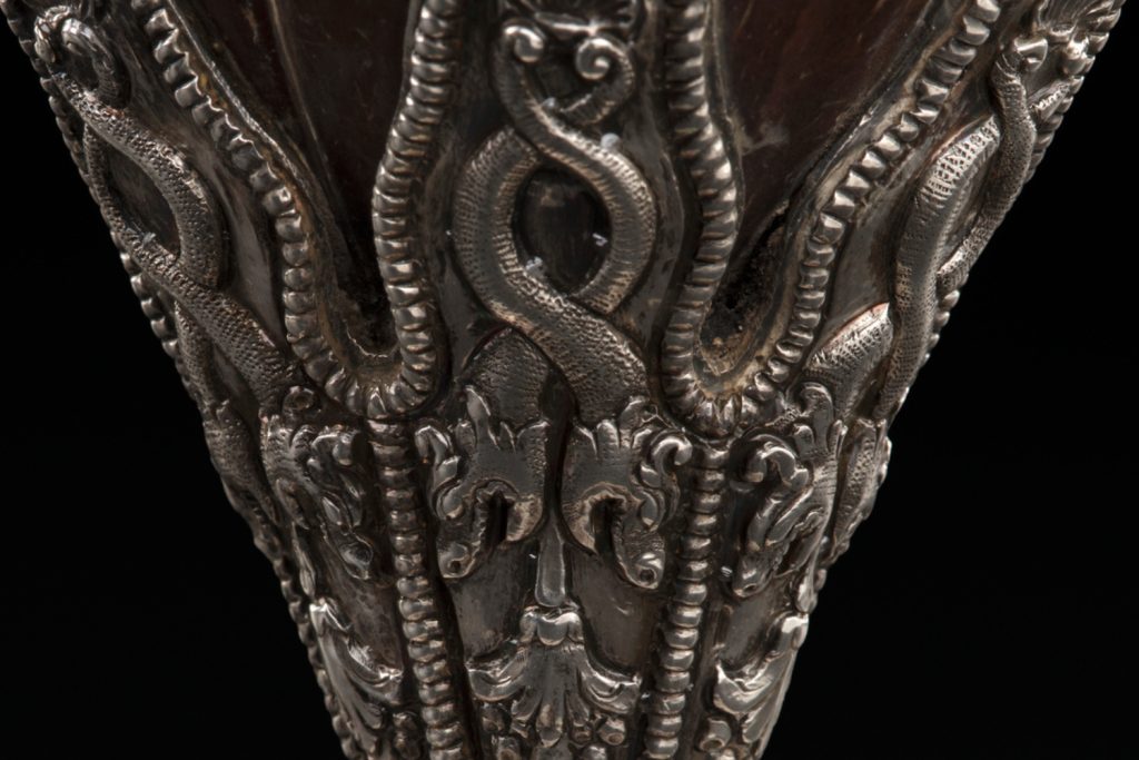 Closeup of the stem of the coconut cup focused on the silver decorations. The silver features intertwined snakes or dragons pointed downward towards the base.