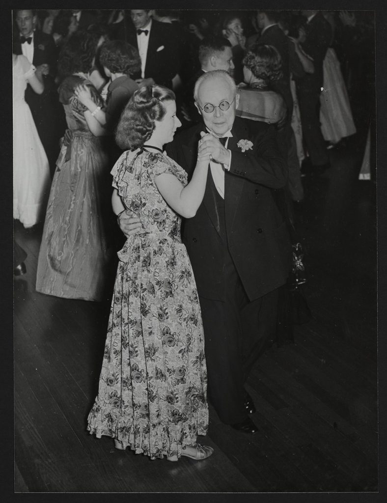 An elderly man in a tuxedo and round eyeglasses dances formally with a young woman in a floral dress. Behind them are half a dozen other dancing pairs.