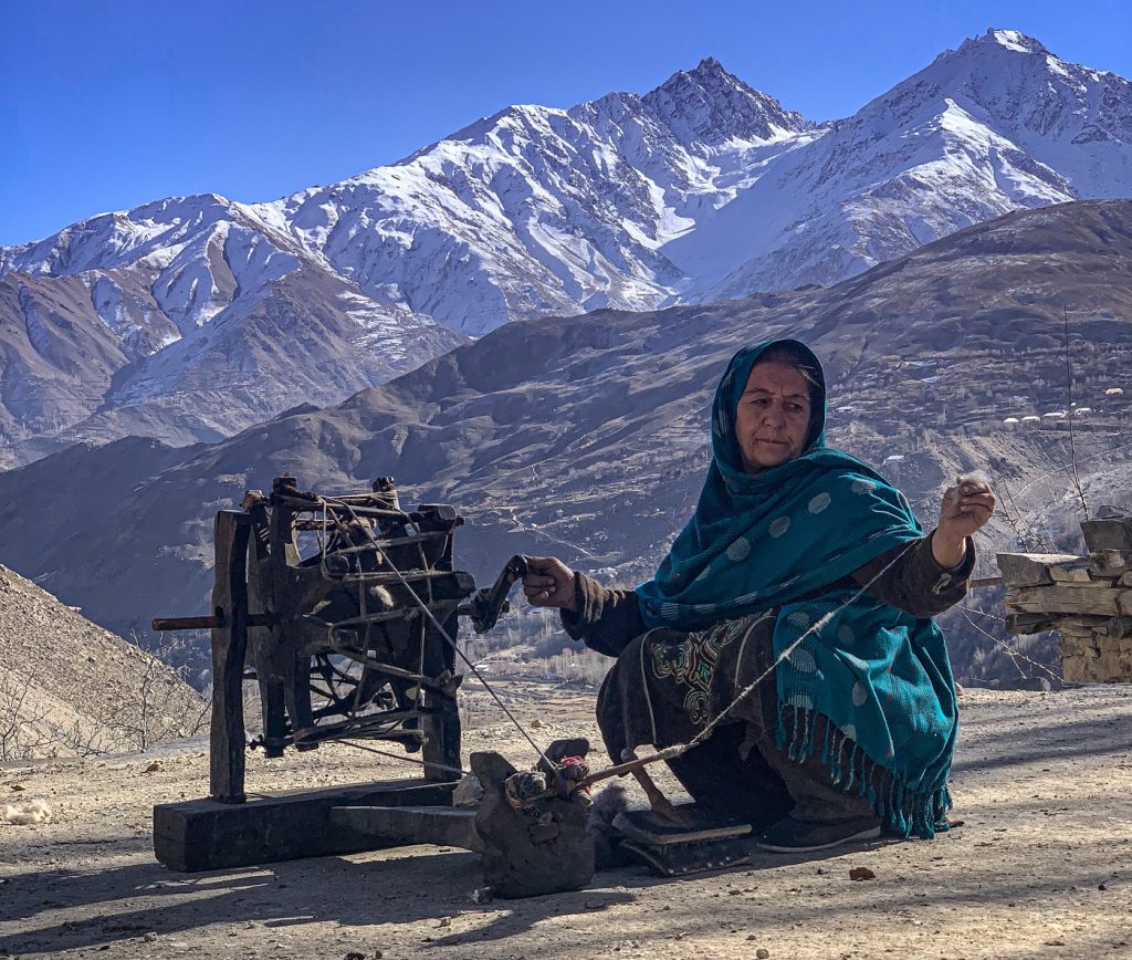 A woman wearing a blue shawl kneels and masterfully pulls a thread through a wooden device outside on a flat, dirt surface. Behind her is an awe-inspiring vista of high, snow-covered mountains.