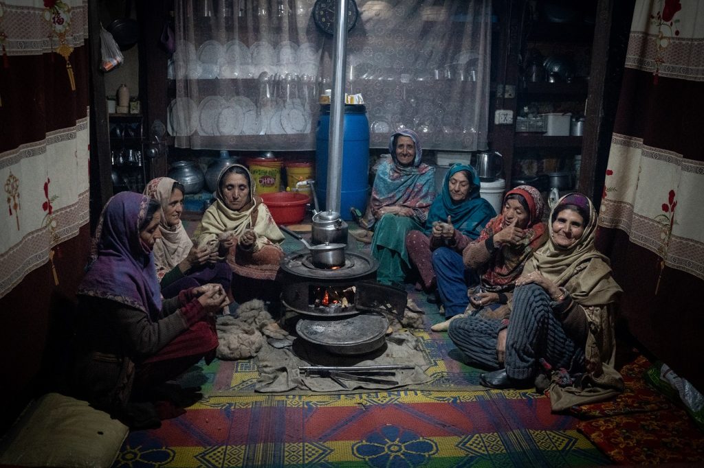 An intimate, softly lit portrait of a group of seven women, all elderly and wearing colourful shawls, happily gathered around a stove inside a home. Some converse, some smile into the camera. Very cosy and clearly a good time had by all.