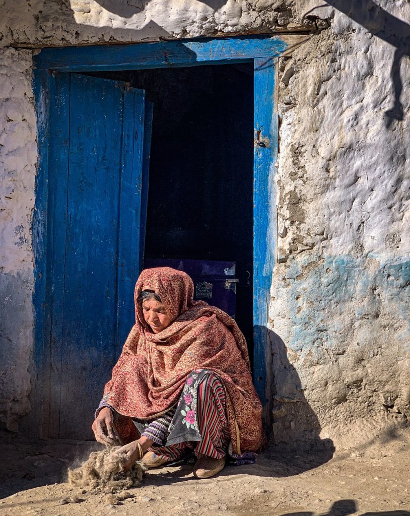 An elderly woman in colourful red and yellow-patterned shawl kneels in front of a building with a blue door. She pours sand into a pile of fibres with a considered look on her face.