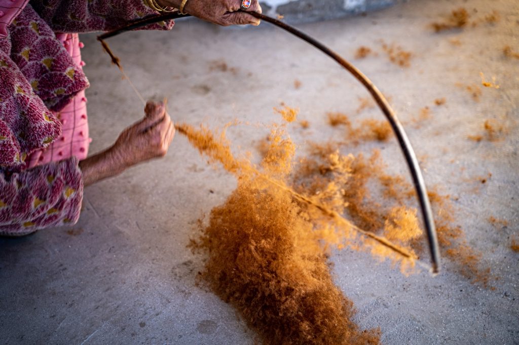 Soft focus closeup of a woman holding a wooden carding bow, shaped like an archery bow. Gold-coloured fibres cling to the bow's string, with a pile accumulating on the dirt ground. 