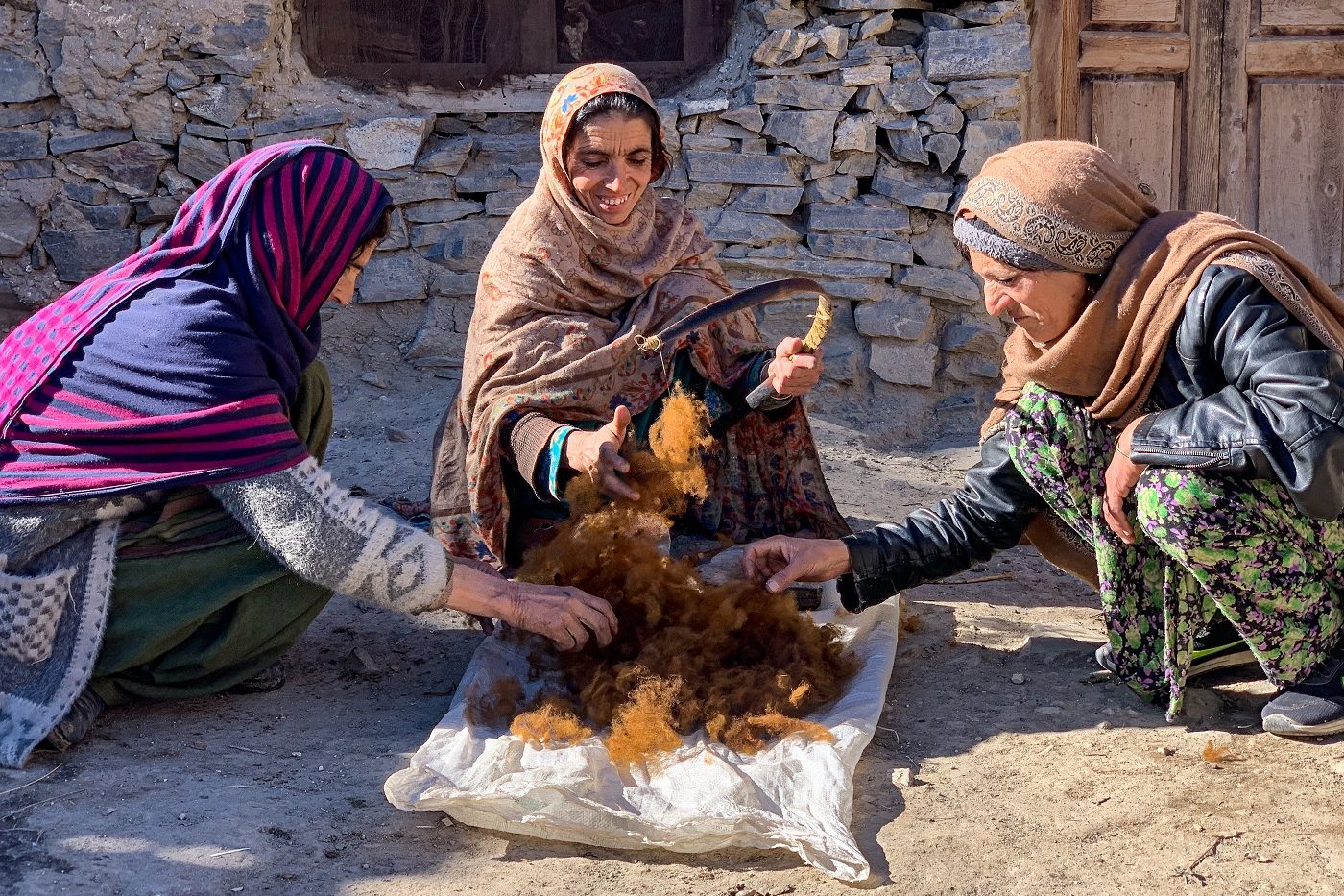 Three elderly women kneel outside a home around a pile of brown-gold fibres resting atop a white cushion. They pick through the fibres, smiling and talking.