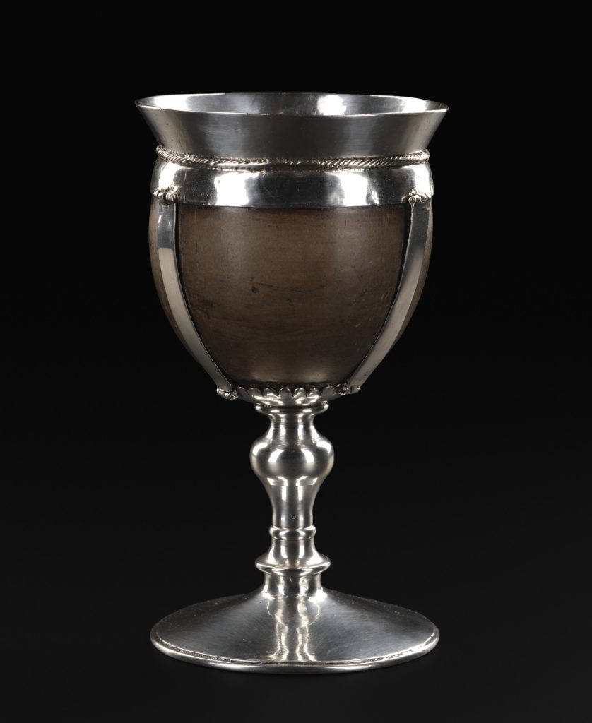 A very smooth, dark brown coconut shell harnessed into a cup with silver straps, a convex silver rim, and plain silver stem, set against a black background.