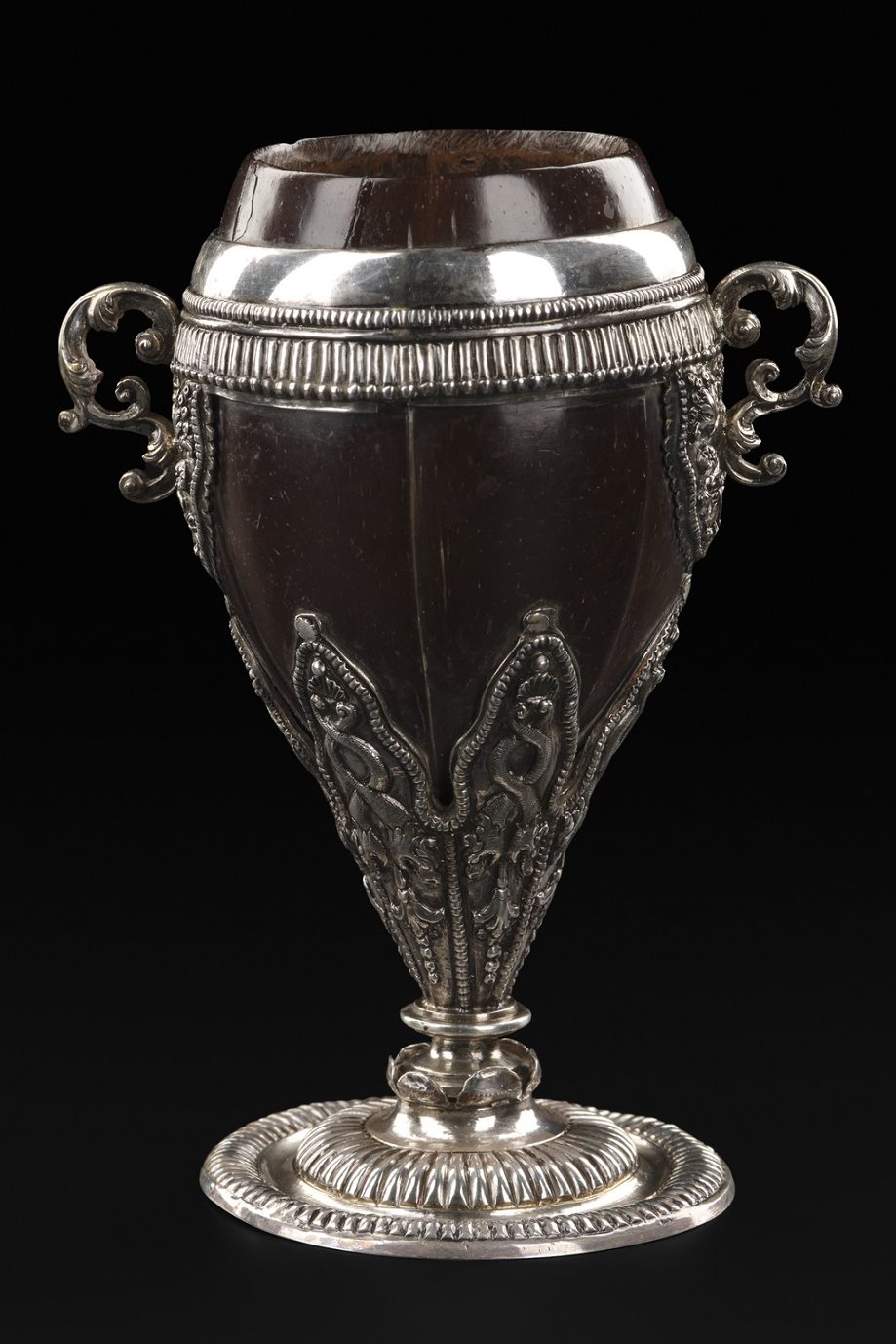 A dark brown polished coconut shell made into an ornate silver drinking cup. A silver rim is at the top, with a silver handle on each side and ornate silver engravings on the stem.