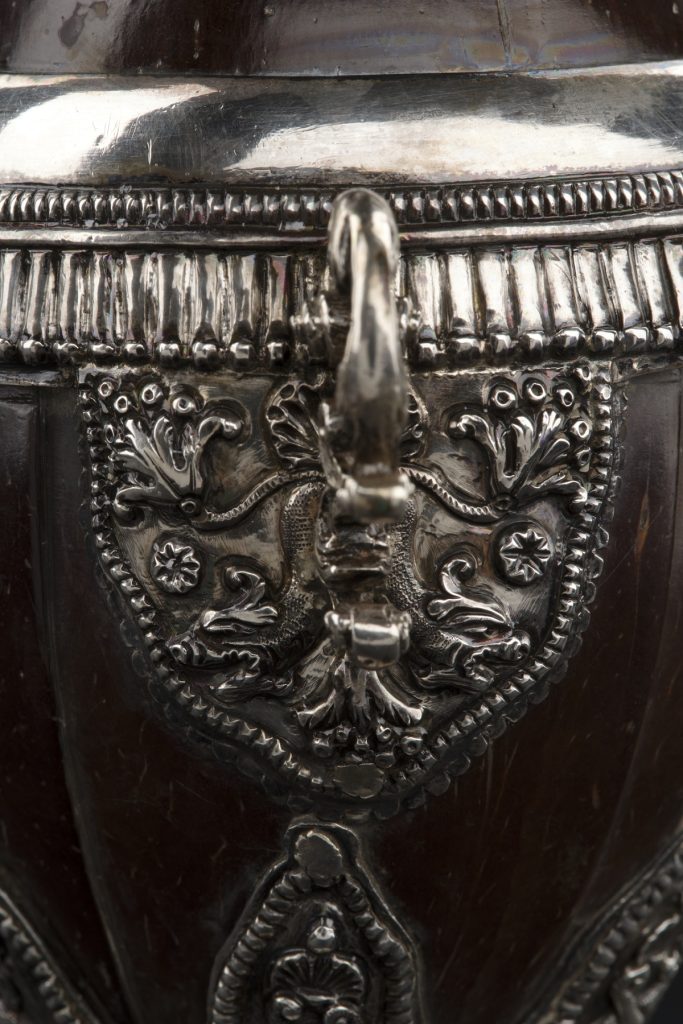 Closeup of an engraved, shield-shaped silver decoration on the dark brown coconut cup. The silver section features flowers resembling fleur-de-lis. 